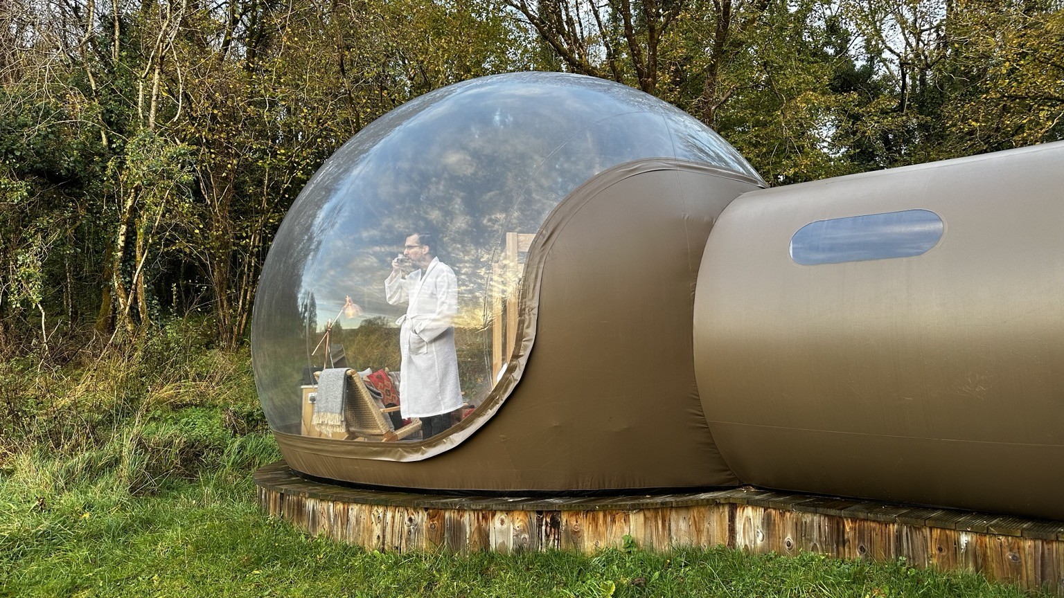 A man in a bathrobe, standing in an inflatable dome tent in the woods